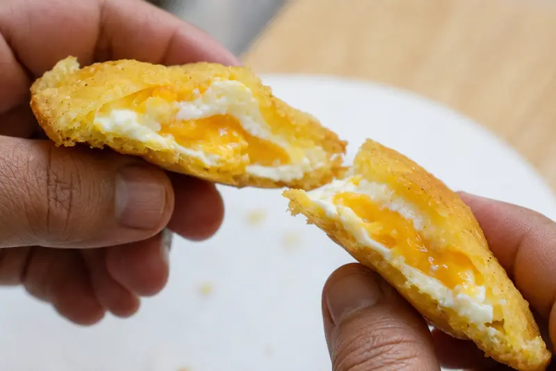 A fried arepa de huevo broken open in two hands, the cooked egg white and soft yolk visible inside the golden corn crust