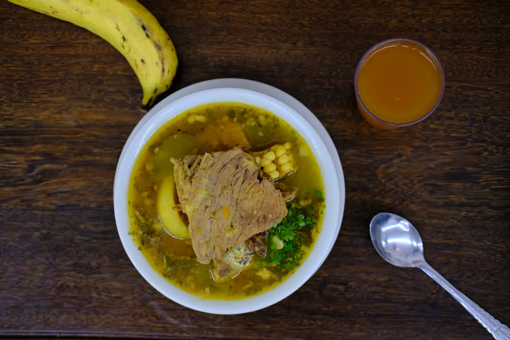 A bowl of traditional Colombian sancocho soup, featuring a piece of meat, corn, and vegetables, served with a banana and a glass of juice on a wooden table.