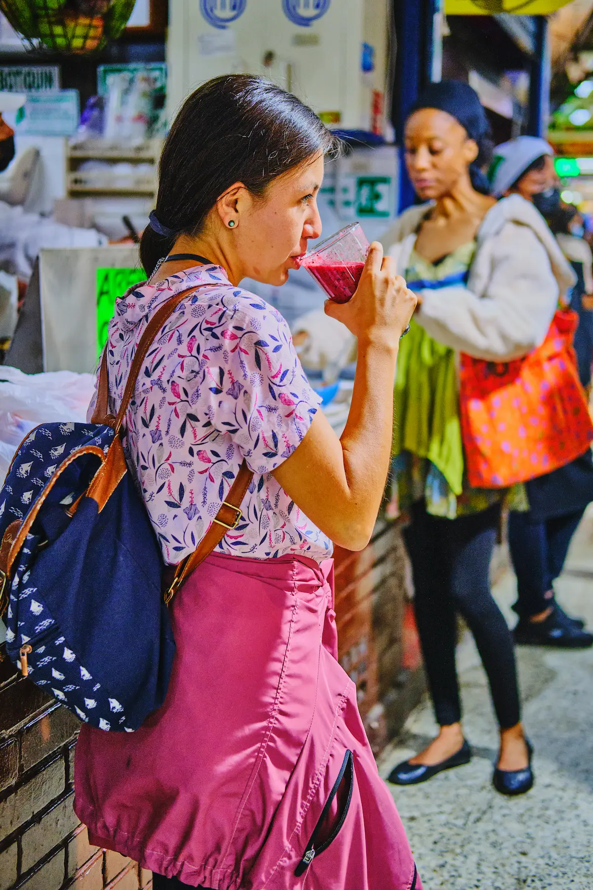 A woman in a floral shirt and pink skirt drinks a red juice at a bustling local market during a food tour in Pasto, Colombia.