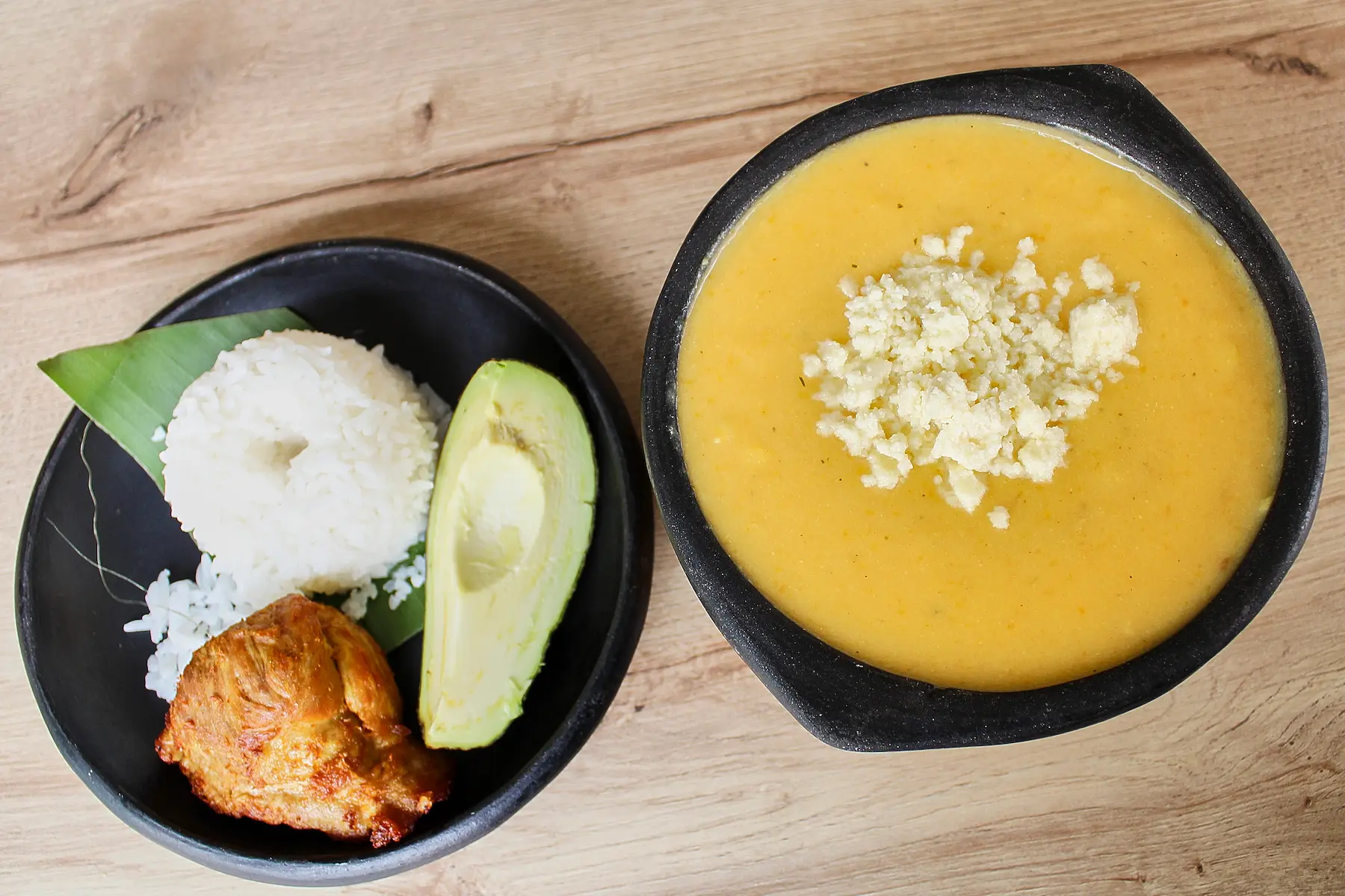 A traditional Colombian meal consisting of a bowl of soup with cheese, served alongside rice, avocado, and a piece of roasted chicken on a black plate, displayed on a wooden table.