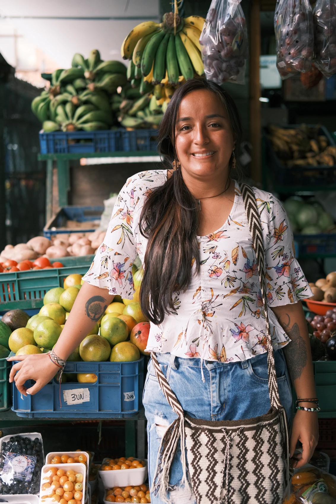 Female fruit market tour guide in floral top smiles while standing next to crates of colorful fruits including bananas, limes, and other tropical produce