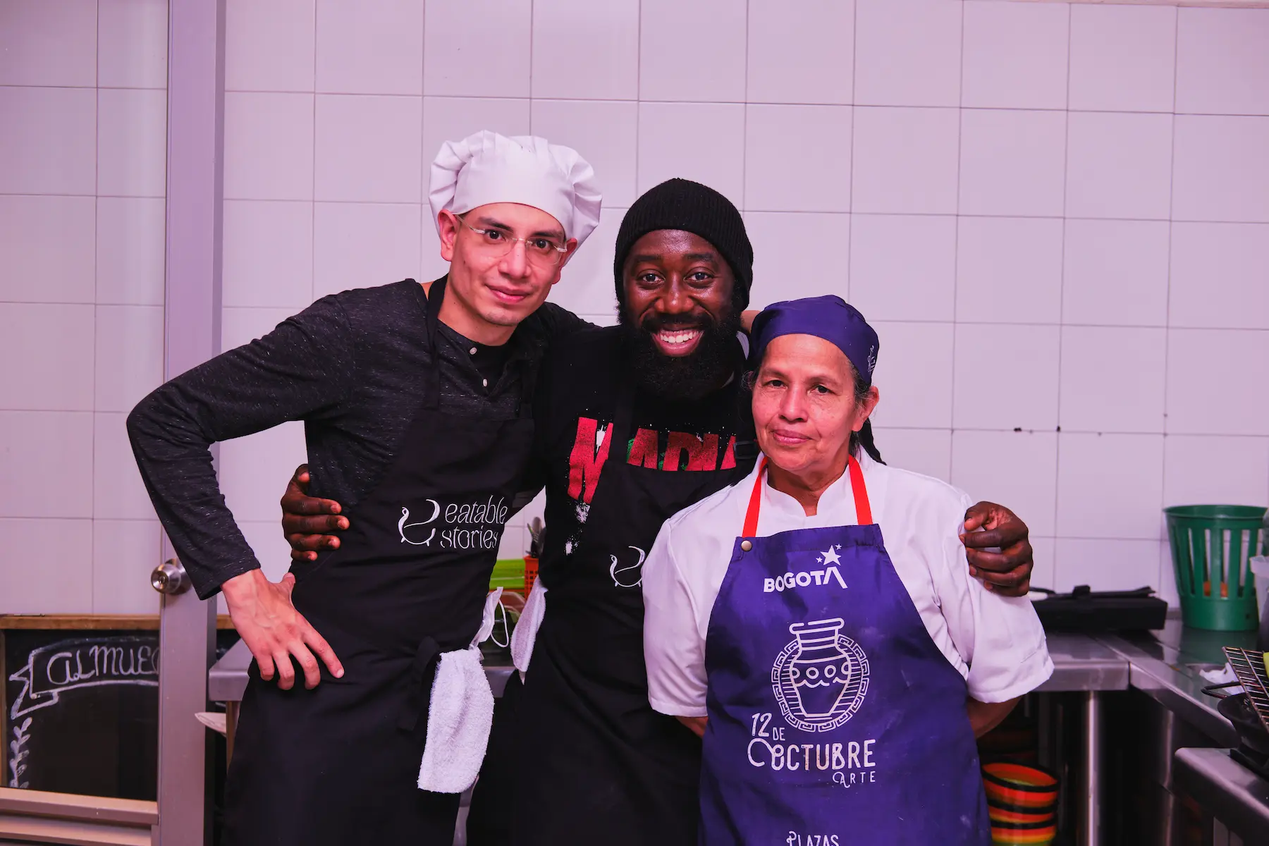 Three chefs of different ethnicities smiling together in a kitchen, prepared to lead a Colombian seafood cooking class. One wears a white chef's hat, another a black cap, and the third a purple Bogota apron.