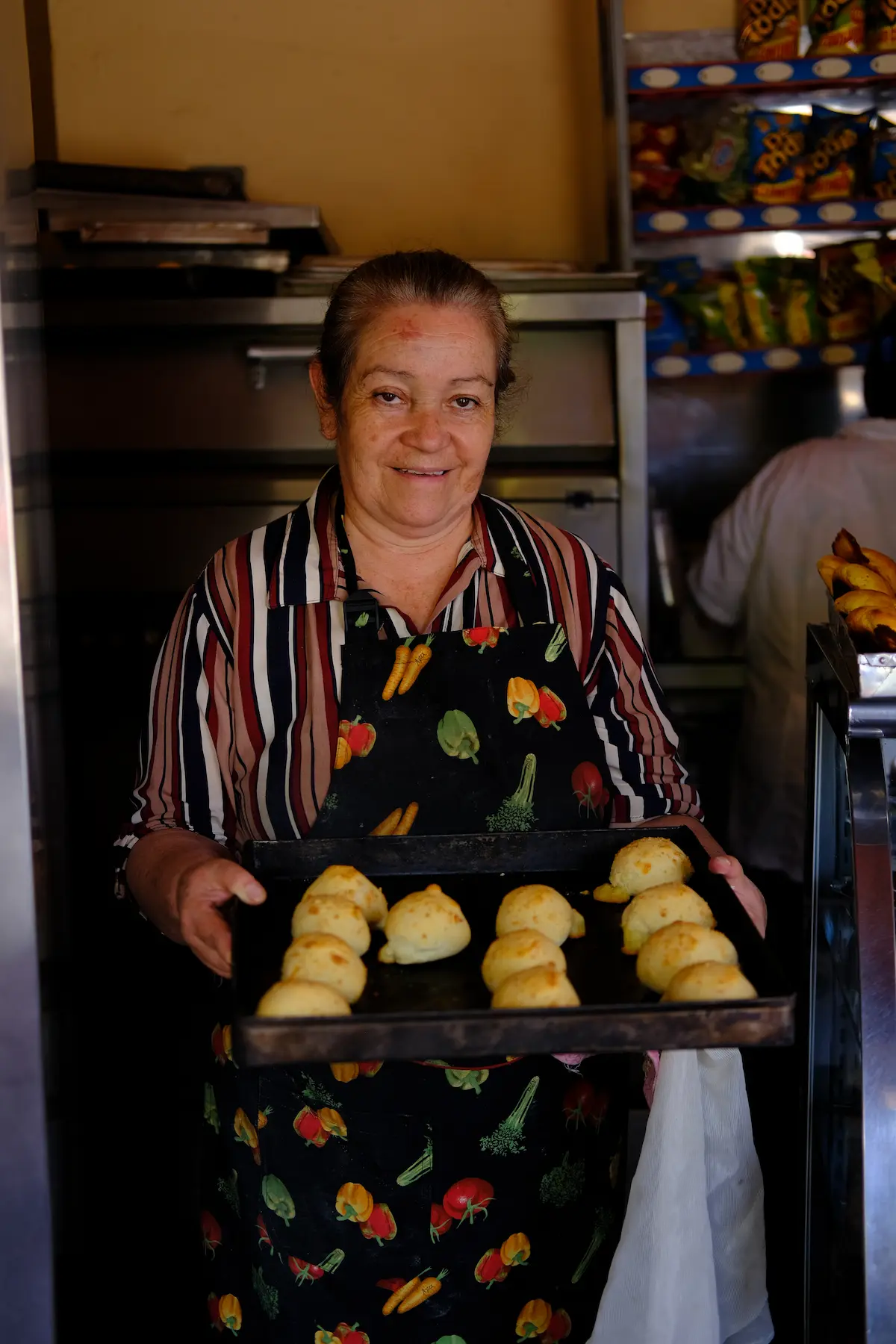 A woman in a striped shirt and vegetable-patterned apron holding a tray of freshly baked buns in a local bakery in Páramo de Cruz Verde, Colombia.