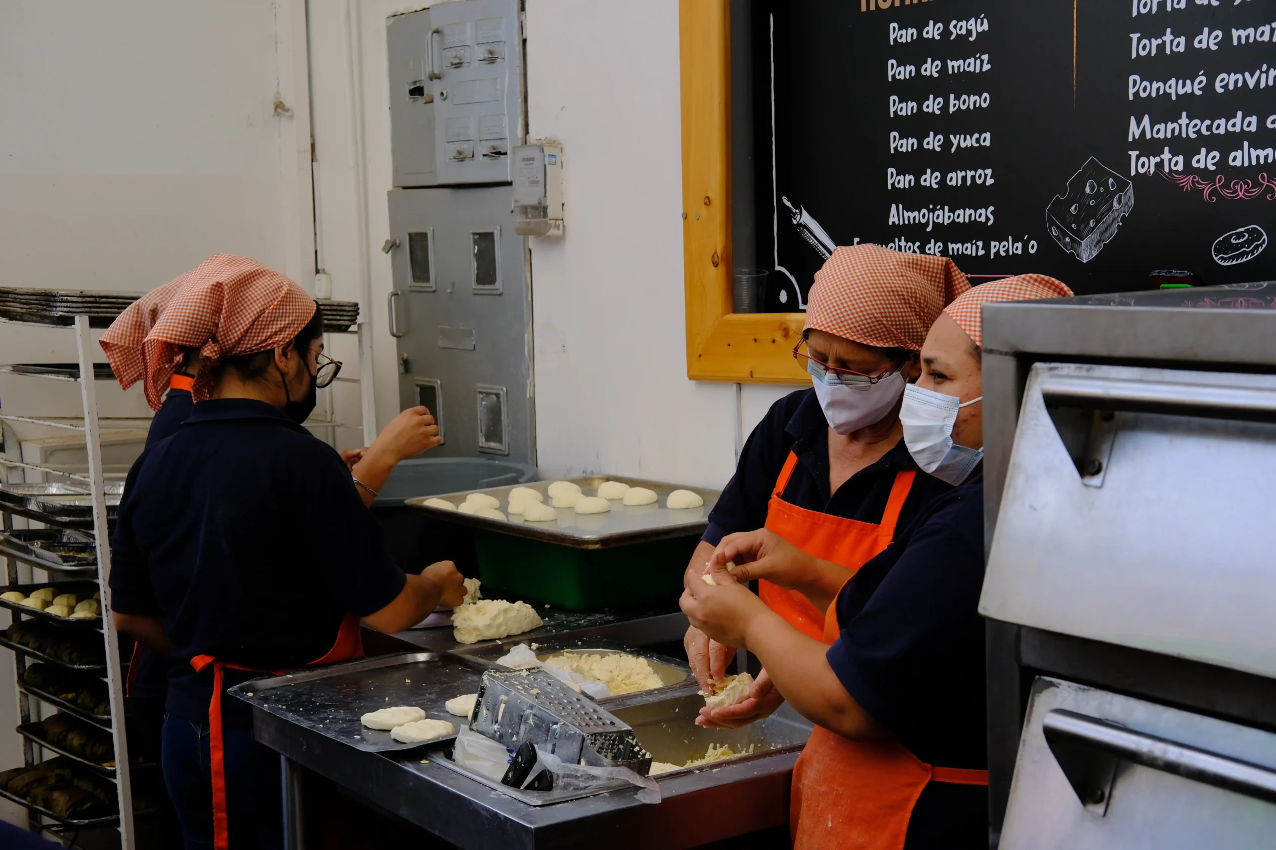 Three women wearing orange aprons and headscarves, working together to prepare dough in a traditional bakery in Páramo de Cruz Verde, Colombia.