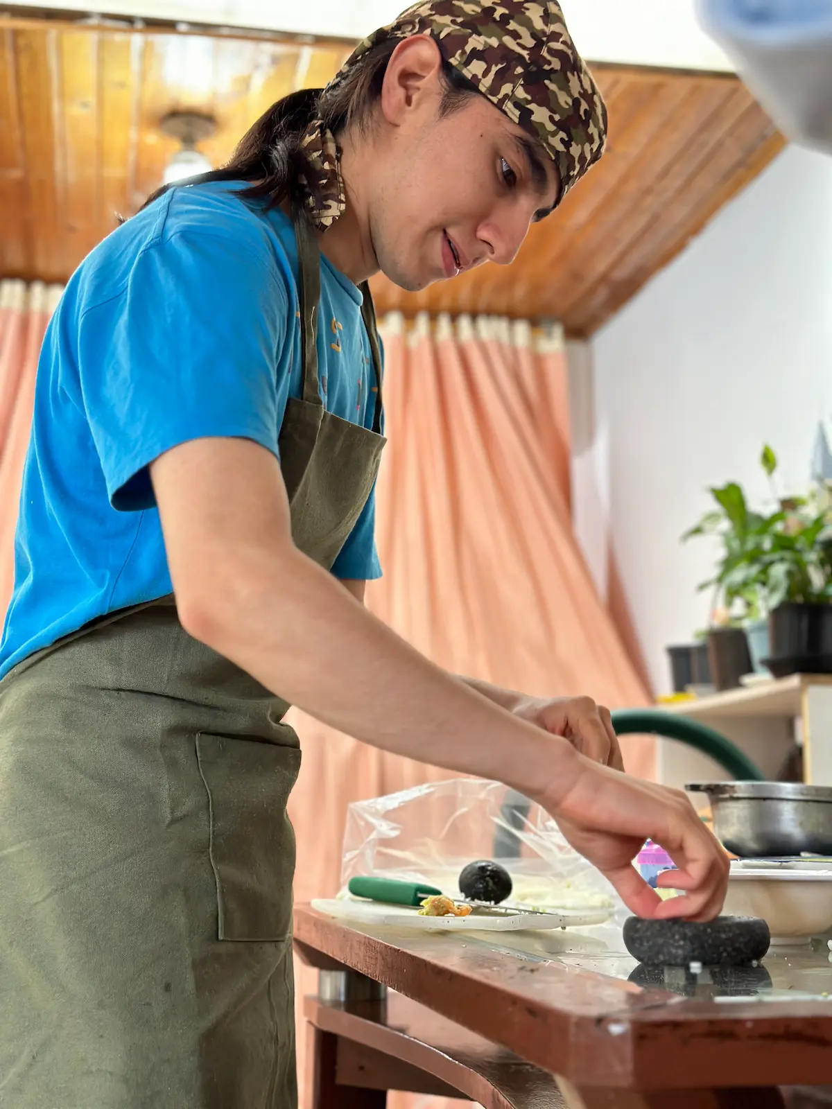 A young man in a blue shirt and camouflage cap prepares ingredients for carimañolas in a home kitchen. He's using a mortar and pestle on a wooden table, with cooking utensils and plastic wrap visible.