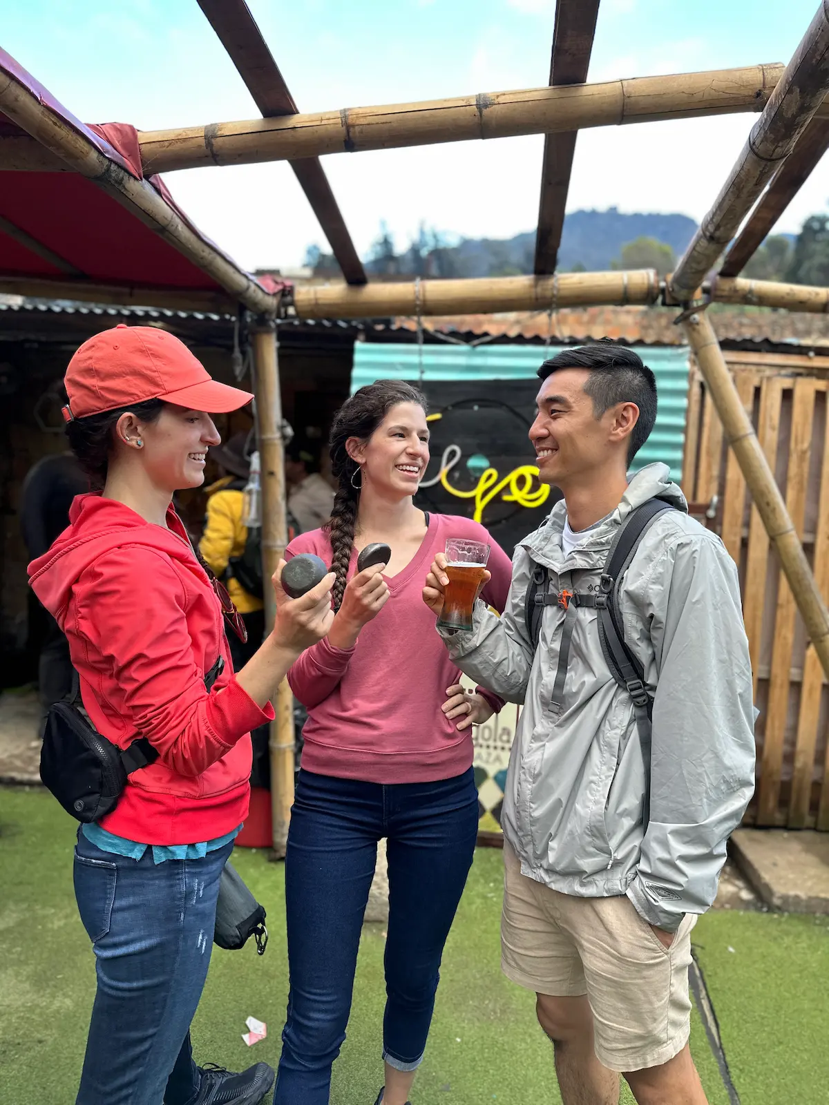 Three diverse friends smiling and chatting under a bamboo structure. One woman in red holds food, another in pink laughs, and a man in gray holds a beer. A neon 'tejo' sign is visible in the background.