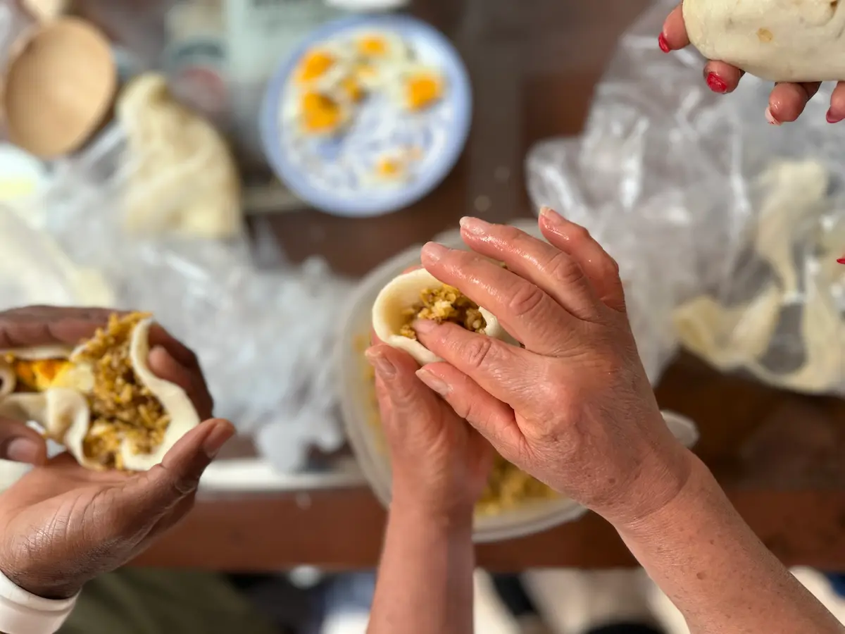 Close-up of hands preparing carimañolas during a Caribbean cooking class. Two people are filling dough pockets with a yellow mixture. A blue plate with finished carimañolas is visible in the background, along with cooking materials on a wooden table.