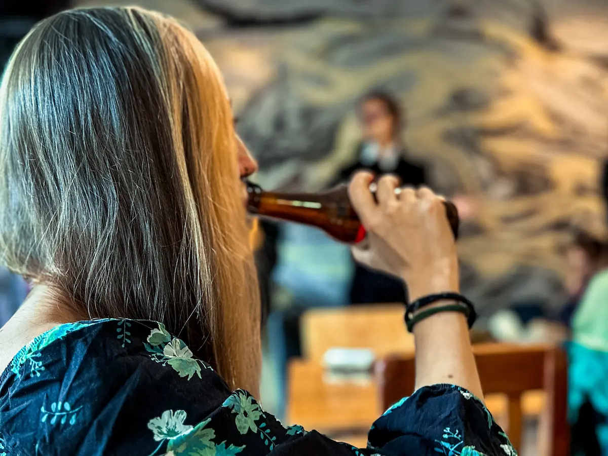 Woman with long blonde hair drinking from a beer bottle, viewed from behind. She's wearing a floral print top. In the background, a blurred mural and other patrons can be seen in what appears to be a restaurant or bar setting.